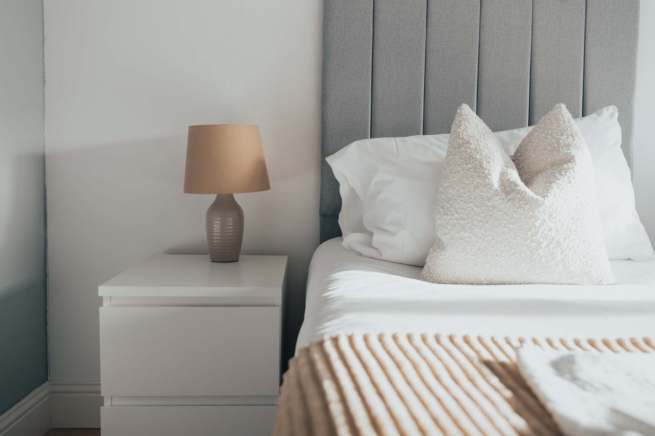 Bedside table and lamp beside a made bed with a textured cushion